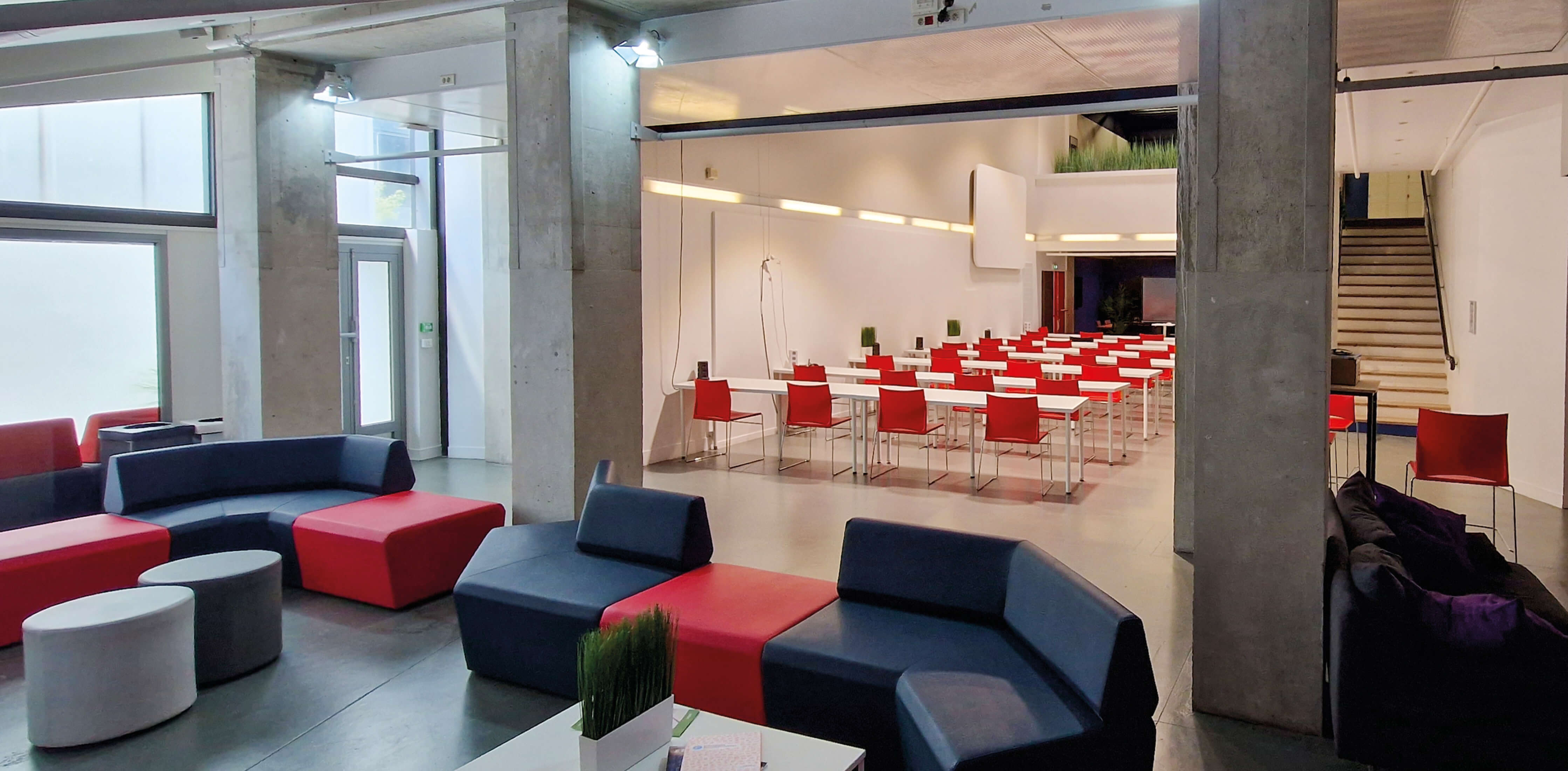 Photo de la salle de classe de l’amphithéâtre de l’École 2600 après reconversion, prise depuis l’espace détente aménagé avec des sièges et canapés bleus, rouges et gris.