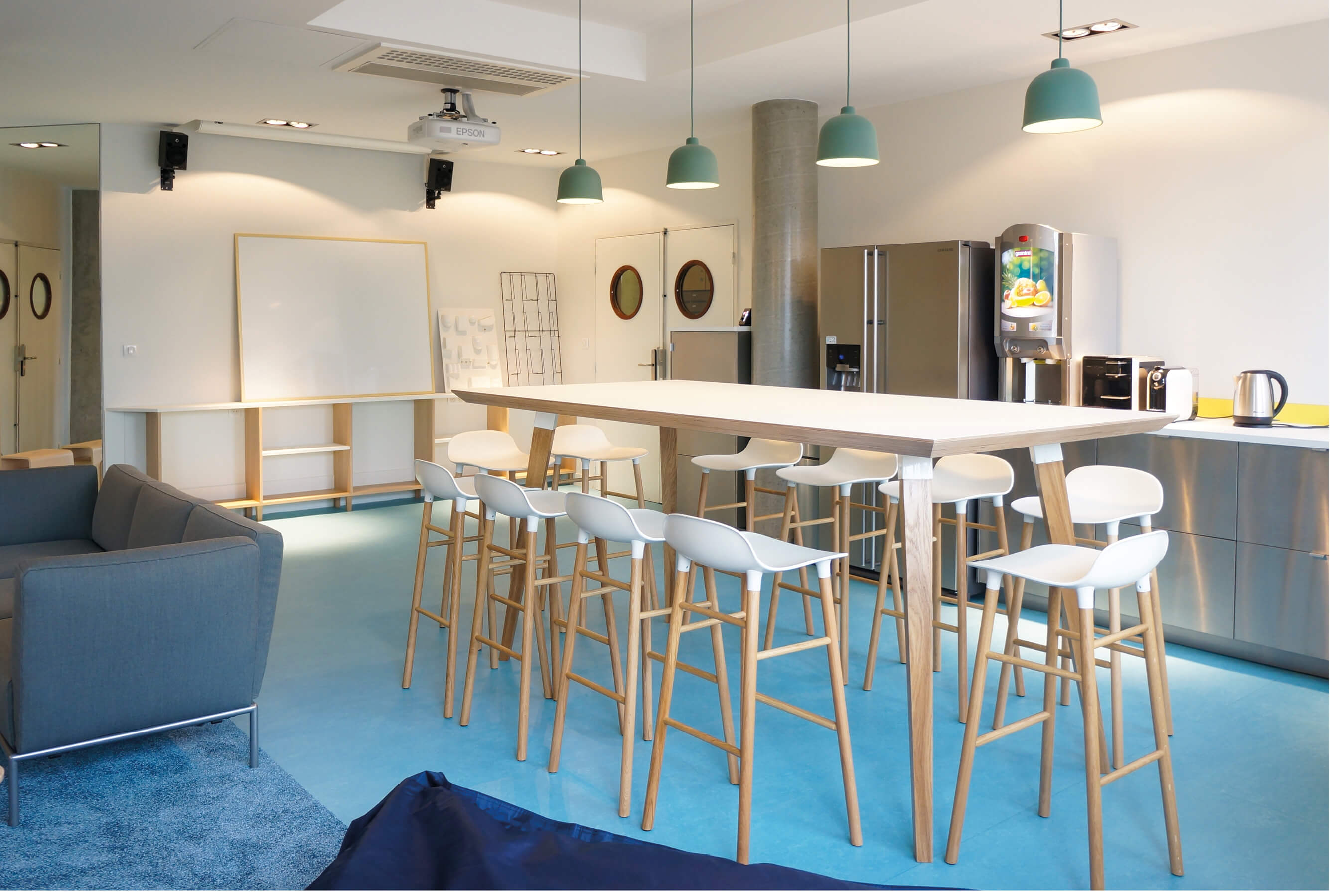 Photo de la salle des professeurs de l’Essec avec la table haute au premier plan, le coin cuisine, l’entrée et le mur interactif apparaissent en arrière-plan