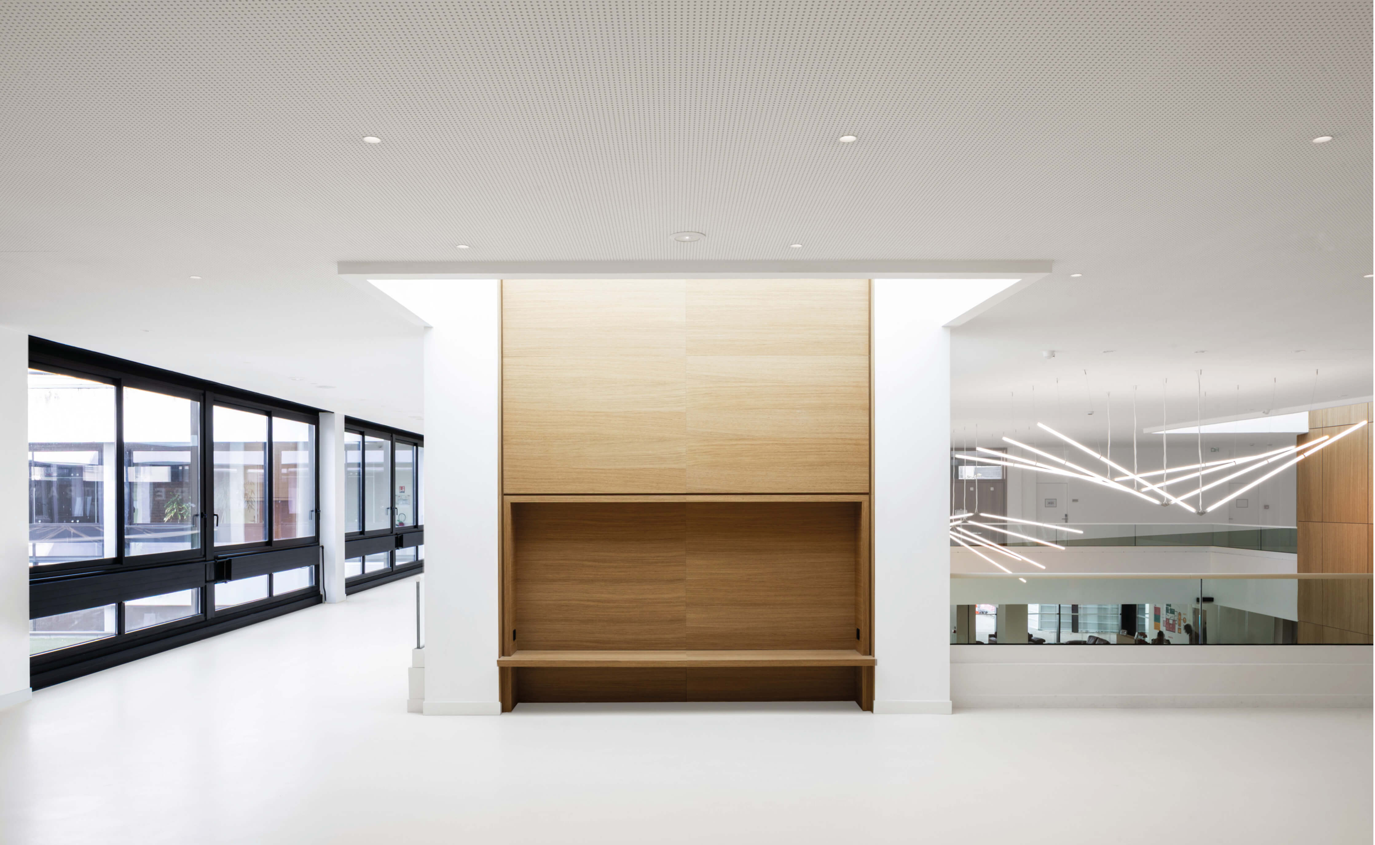 Photo du premier étage du grand hall de l'Essec après la restructuration campus qui laisse voir l'alcôve abritant une banquette en bois de face, les fenêtres et le grand lustre de Simon Deschamps de Superlux.