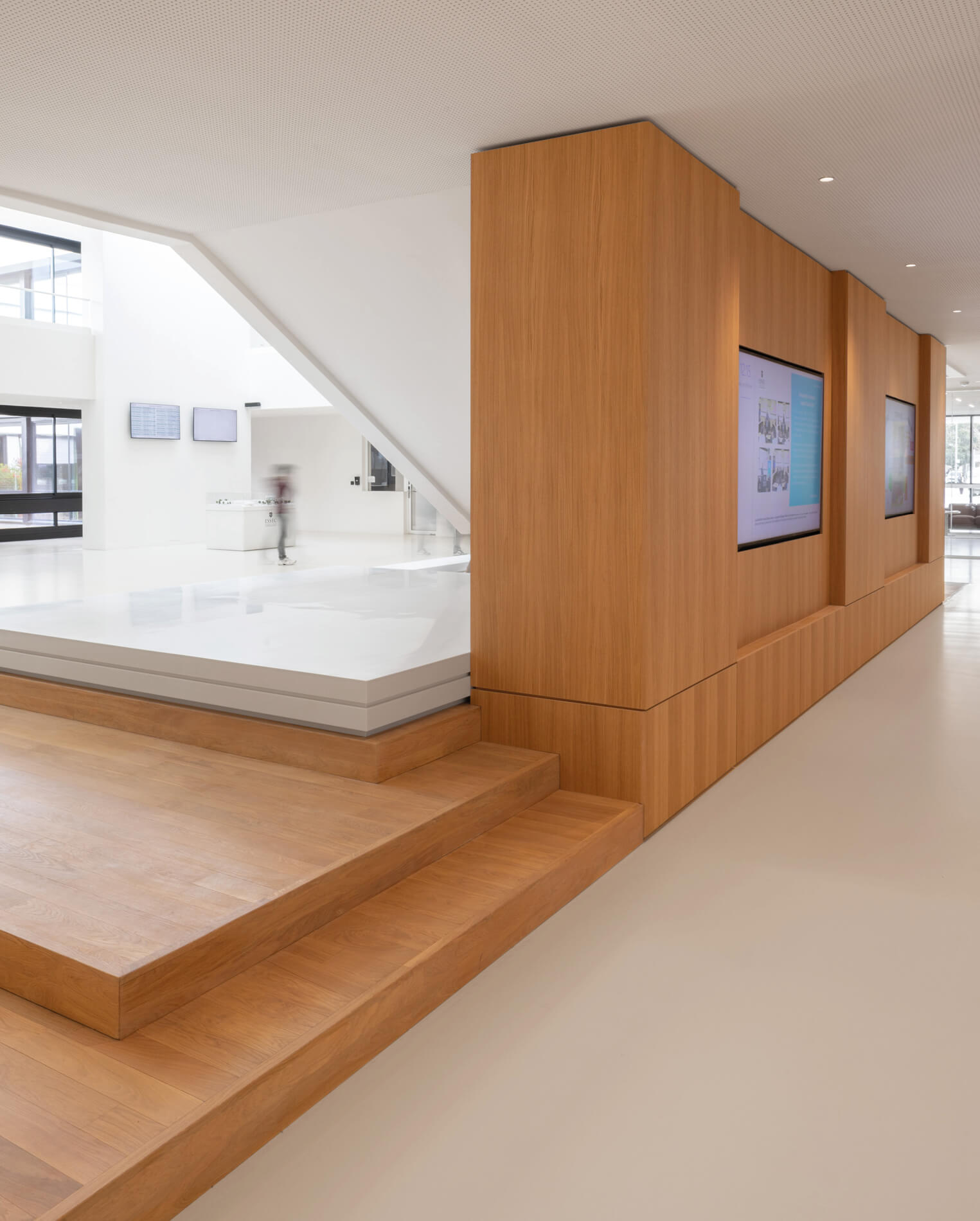 Photo de l'espace sous l'escalier du grand hall de l'Essec. Après les travaux de restructuration campus, il abrite une partie des gradins et un couloir avec des écrans.