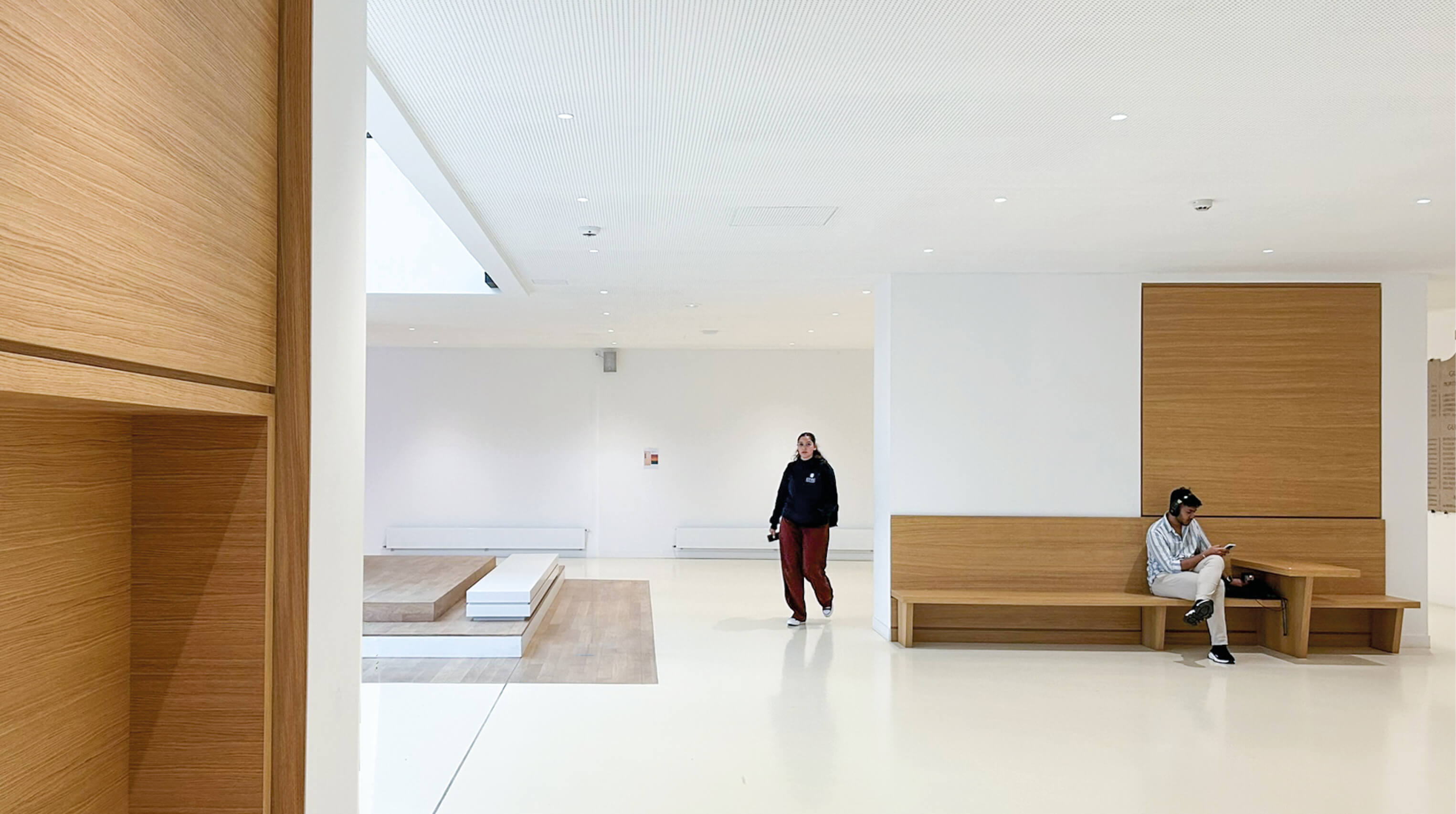 Photo lumineuse du premier étage du grand hall de l'essec avec une élève qui marche, un autre assis sur une banquette en bois.