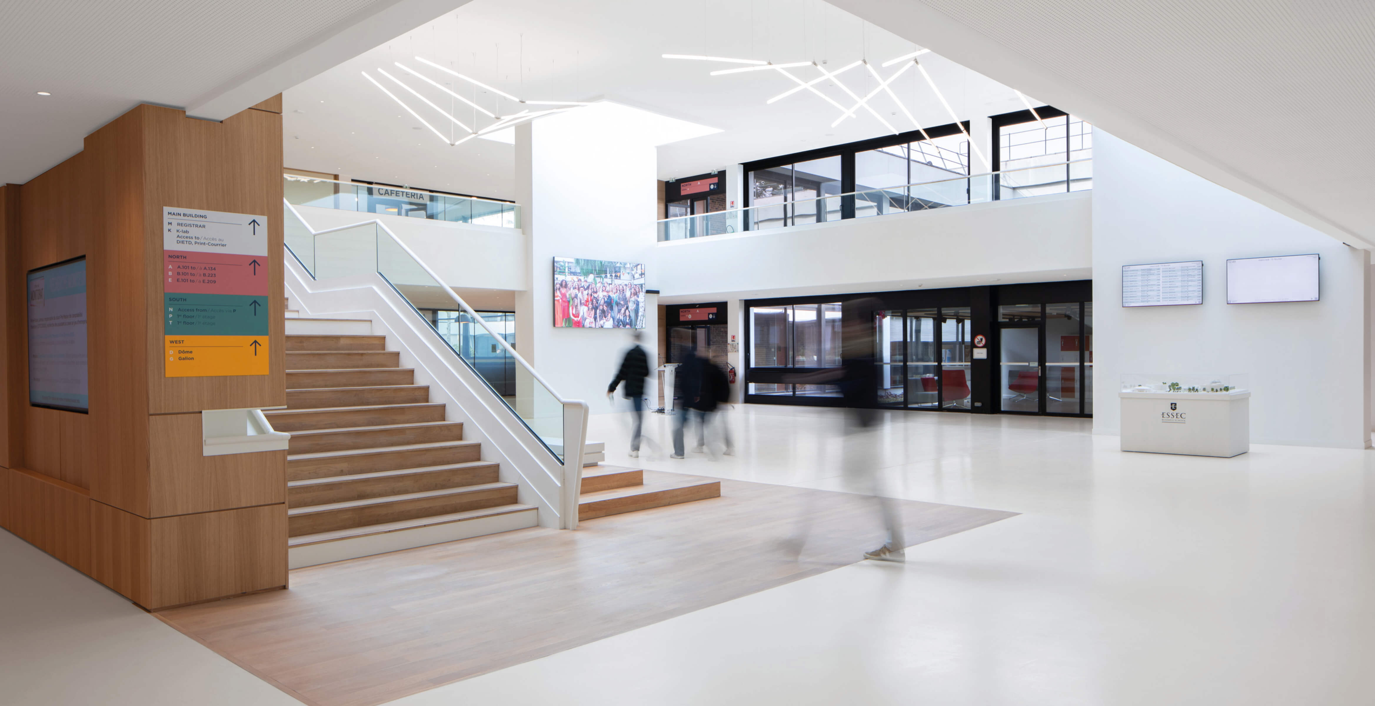 Photo en plan large du grand hall du l'Essec après restructuration du campus avec le bas de l'escalier en premier plan, la grand salle parcourue par des silhouettes et les portes d'entrée dans le fond.
