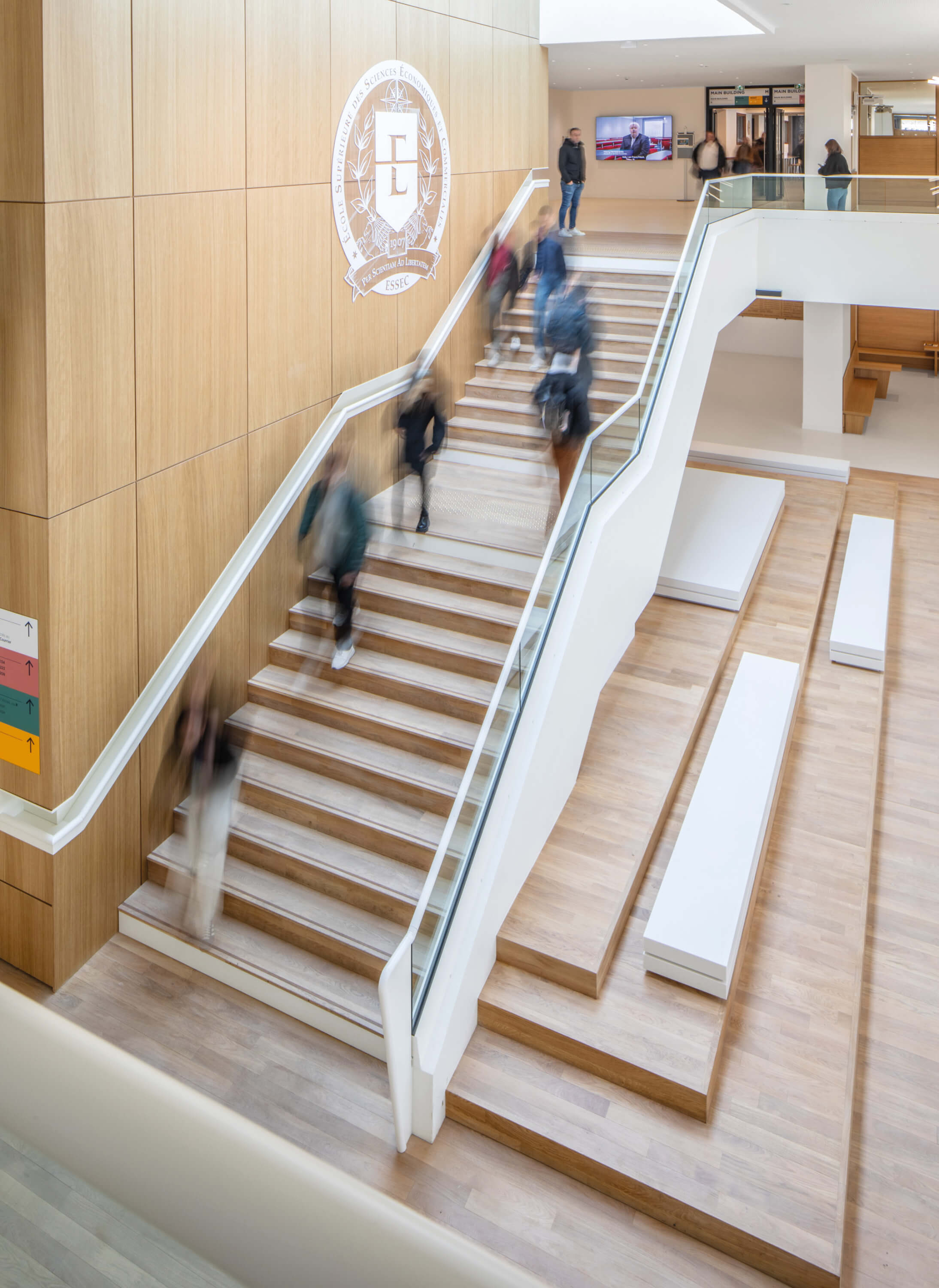 Photo de l'escalier pris du premier étage du grand hall avec des silhouettes l'empruntant après le projet de restructuration du campus de l'Essec.
