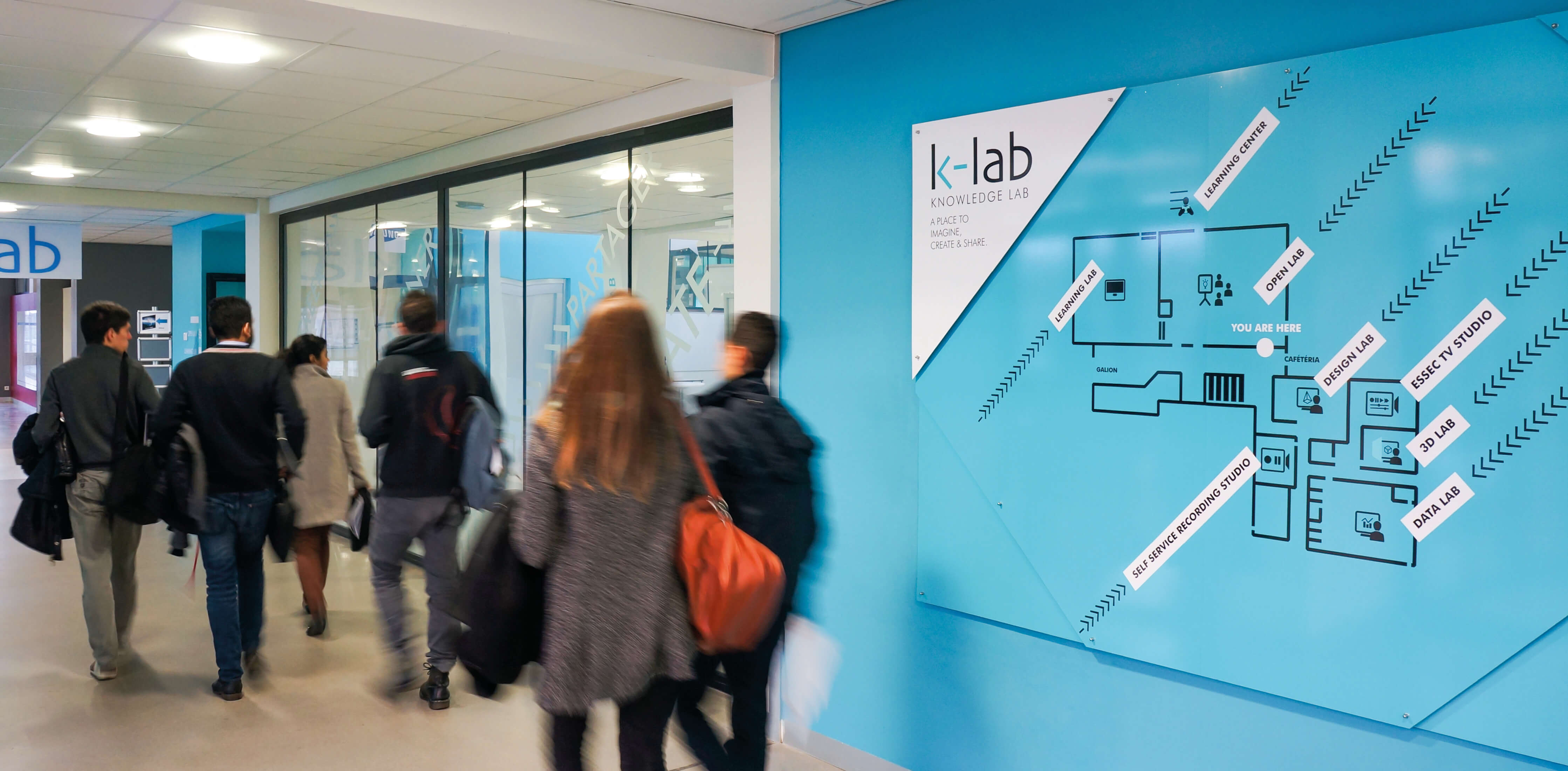 Groupe d’élèves passant devant le mur bleu signalant l’espace K-Lab de l’Essec avec un plan des différentes zones.