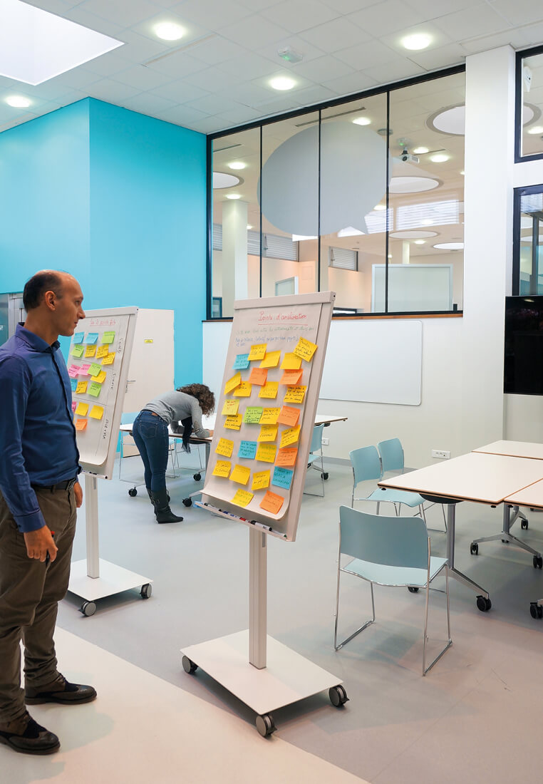 Homme en train de lire les post-its de couleurs laissés sur un pupitre de la salle modulable du K-Lab de l’Essec.