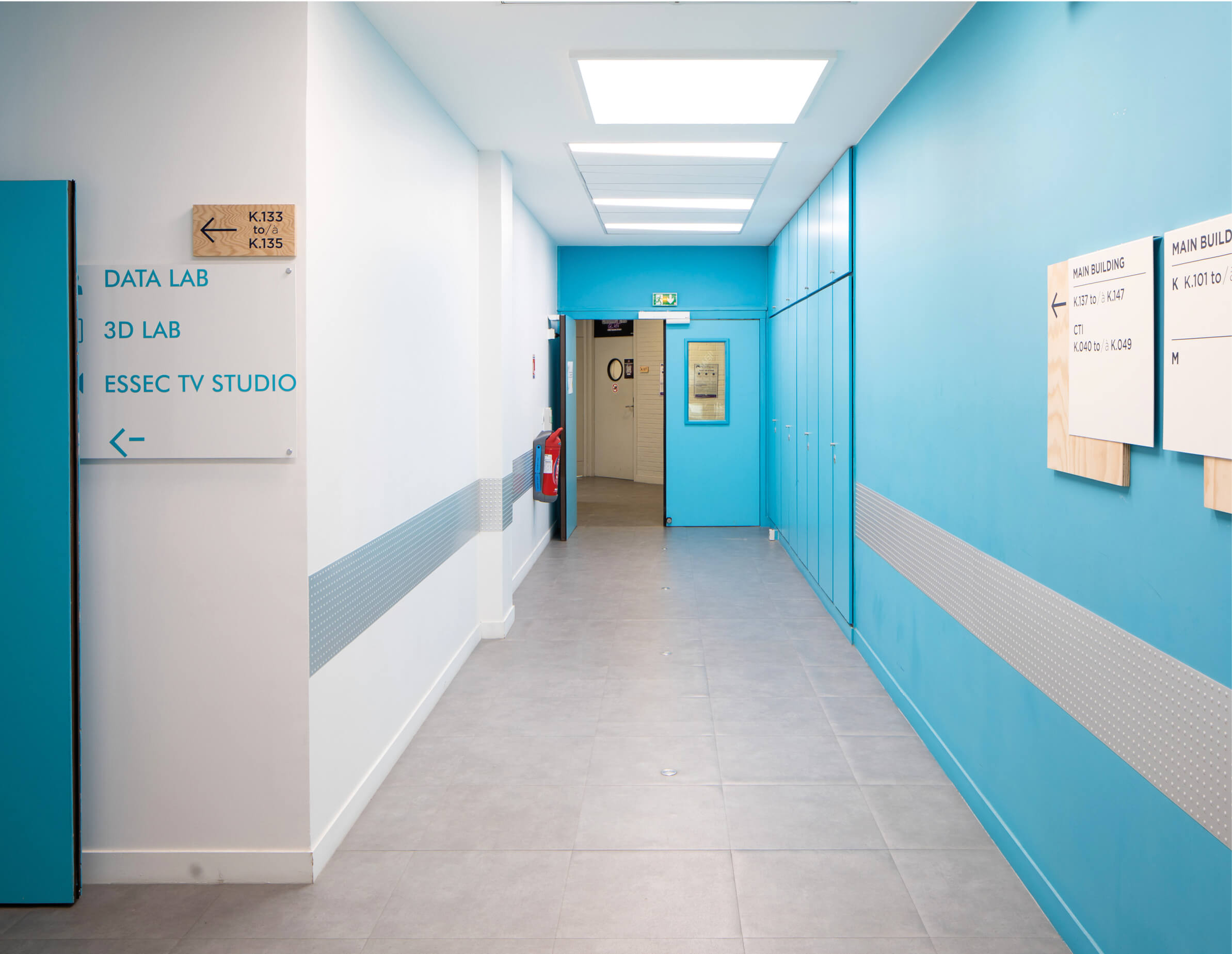 Couloir peint en blanc et bleu avec des signalétiques orientant vers les différents espaces du K-Lab de l’Essec.