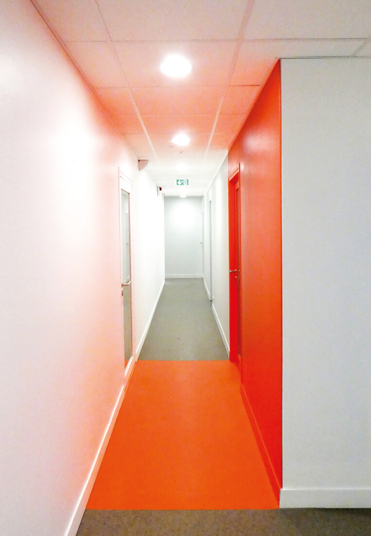 Photo d’un couloir aux murs blancs avec une partie peinte en rouge et, au sol, en orange, le réaménagement du siège de Val d’Oise Habitat La Palette.