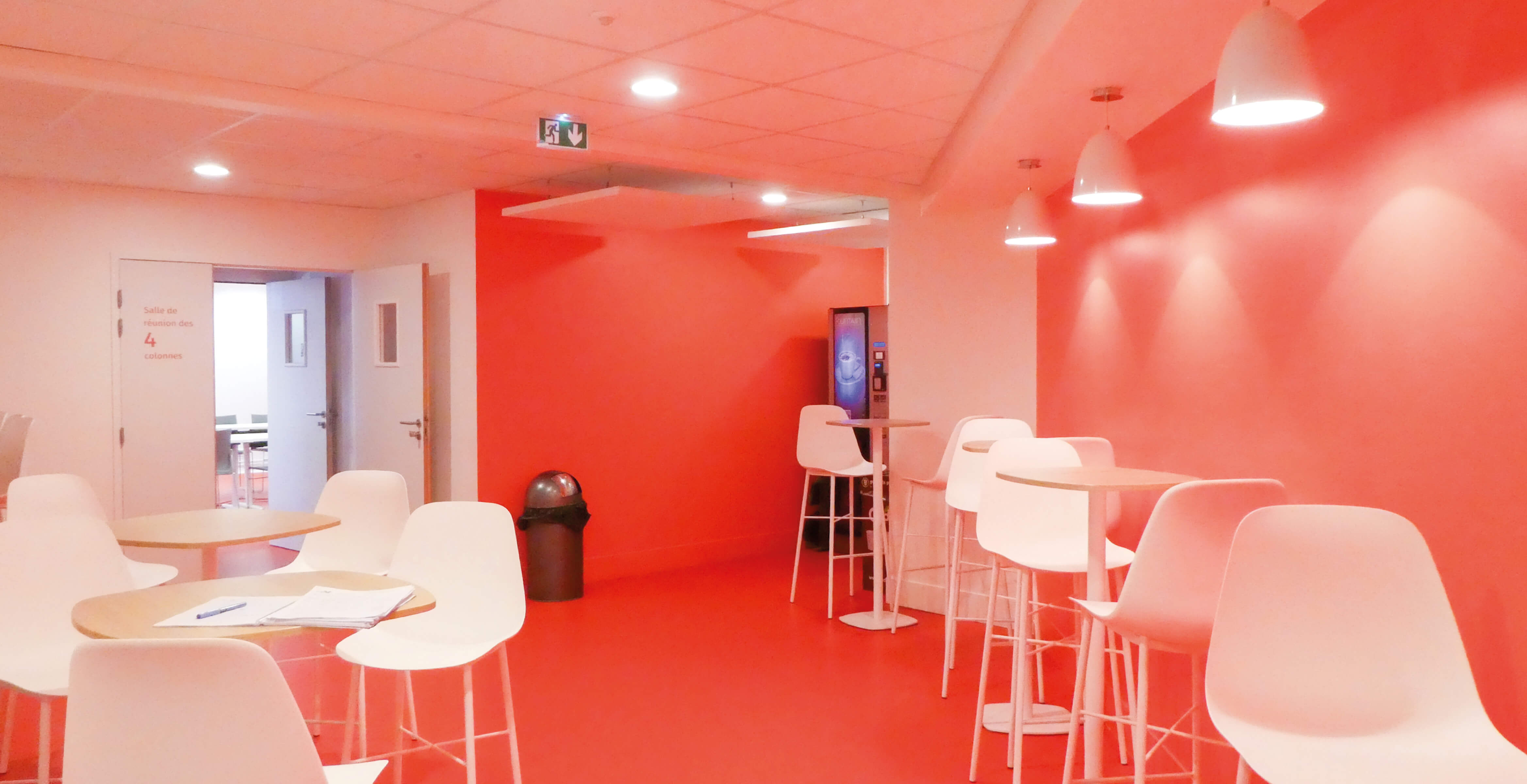 Photo de la salle de repos aux murs rouges avec plusieurs tables hautes au plateau en bois naturel et chaises hautes blanches après le réaménagement du siège de Val d’Oise Habitat La Palette.