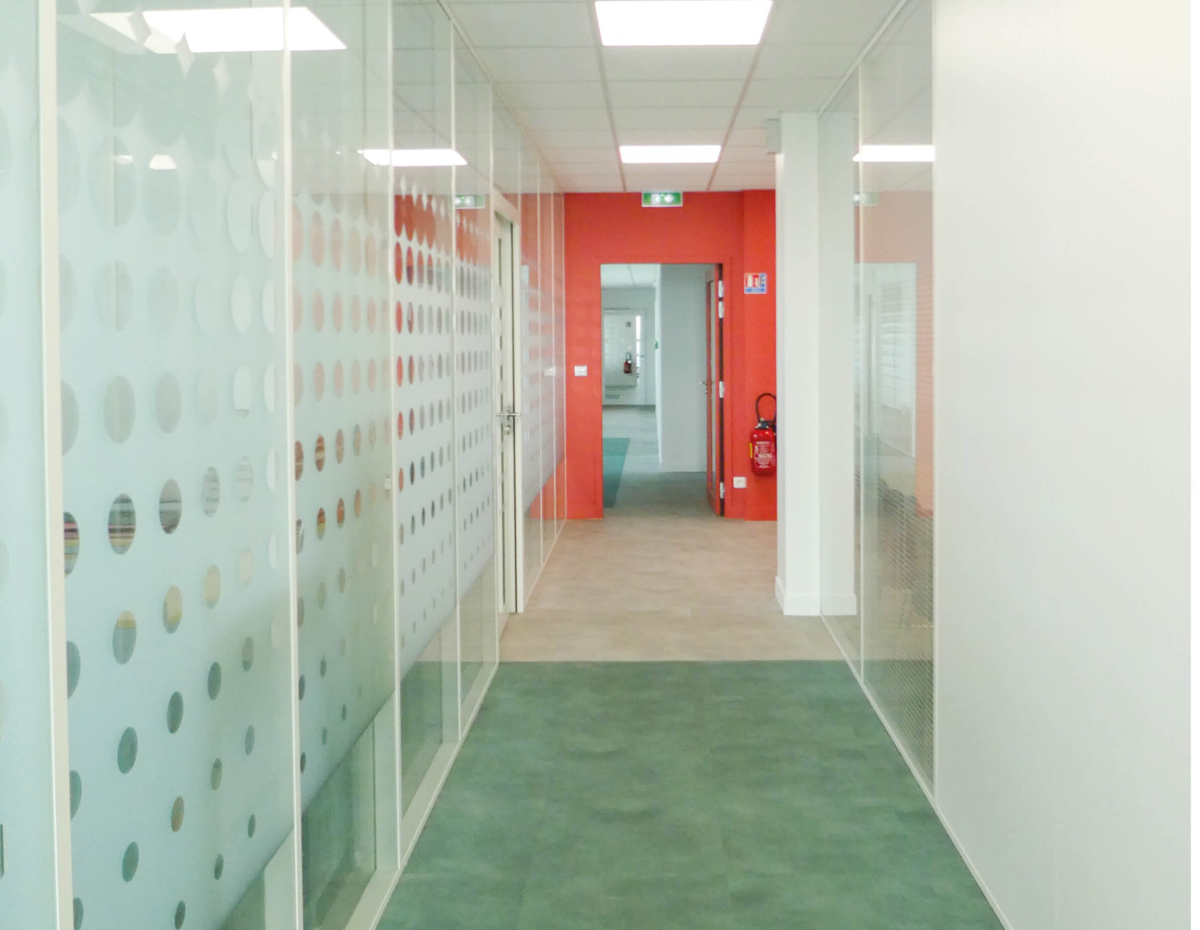 Photo d’un couloir avec une moquette verte, baies vitrées de bureaux et mur rouge en fond après le réaménagement du siège de Val d’Oise Habitat La Palette.