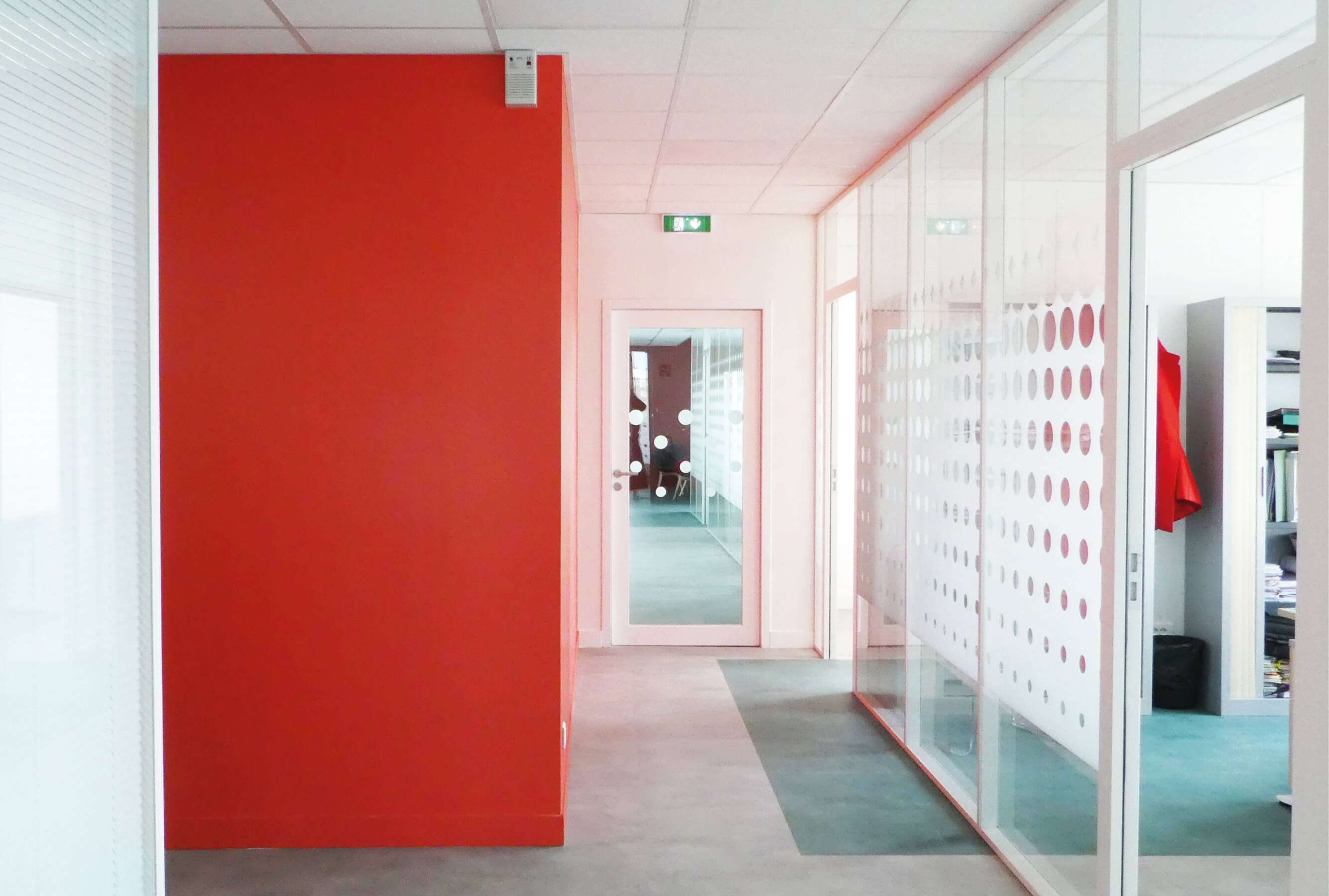 Photo d’un couloir avec moquette verte, mur rouge, bureau avec baie vitrée dont la porte est ouverte après le réaménagement du siège de Val d’Oise Habitat La Palette.