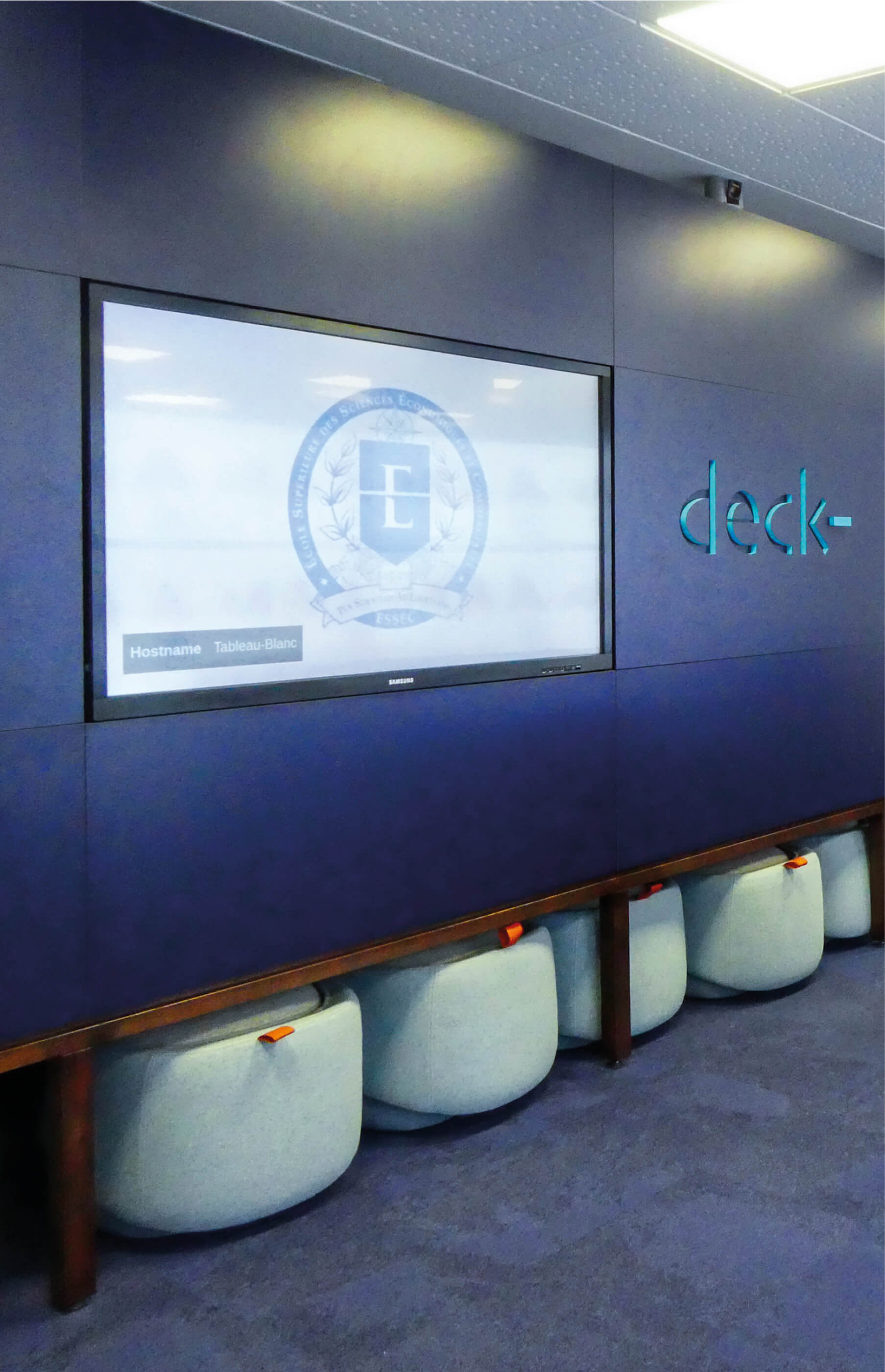 Photo du mur bleu du Deck, la salle virtuelle de l’Essec, avec l’écran du professeur et, au bas, une niche où sont rangés les sièges.