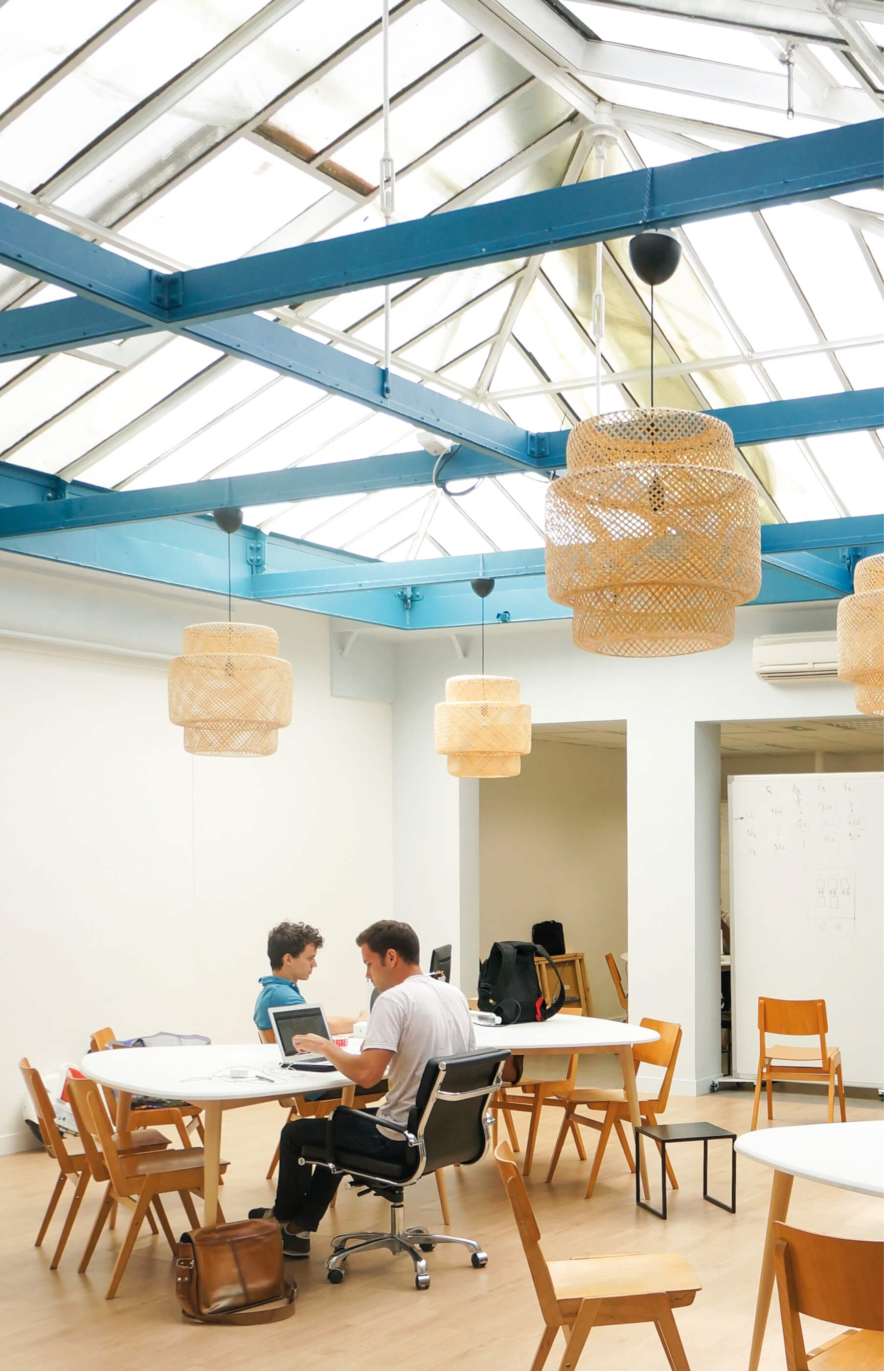 Photo de deux hommes en train de travailler dans le nouvel espace verrière du Schoolab avec poutres peintes en bleu, mobilier en bois, suspensions en osier.