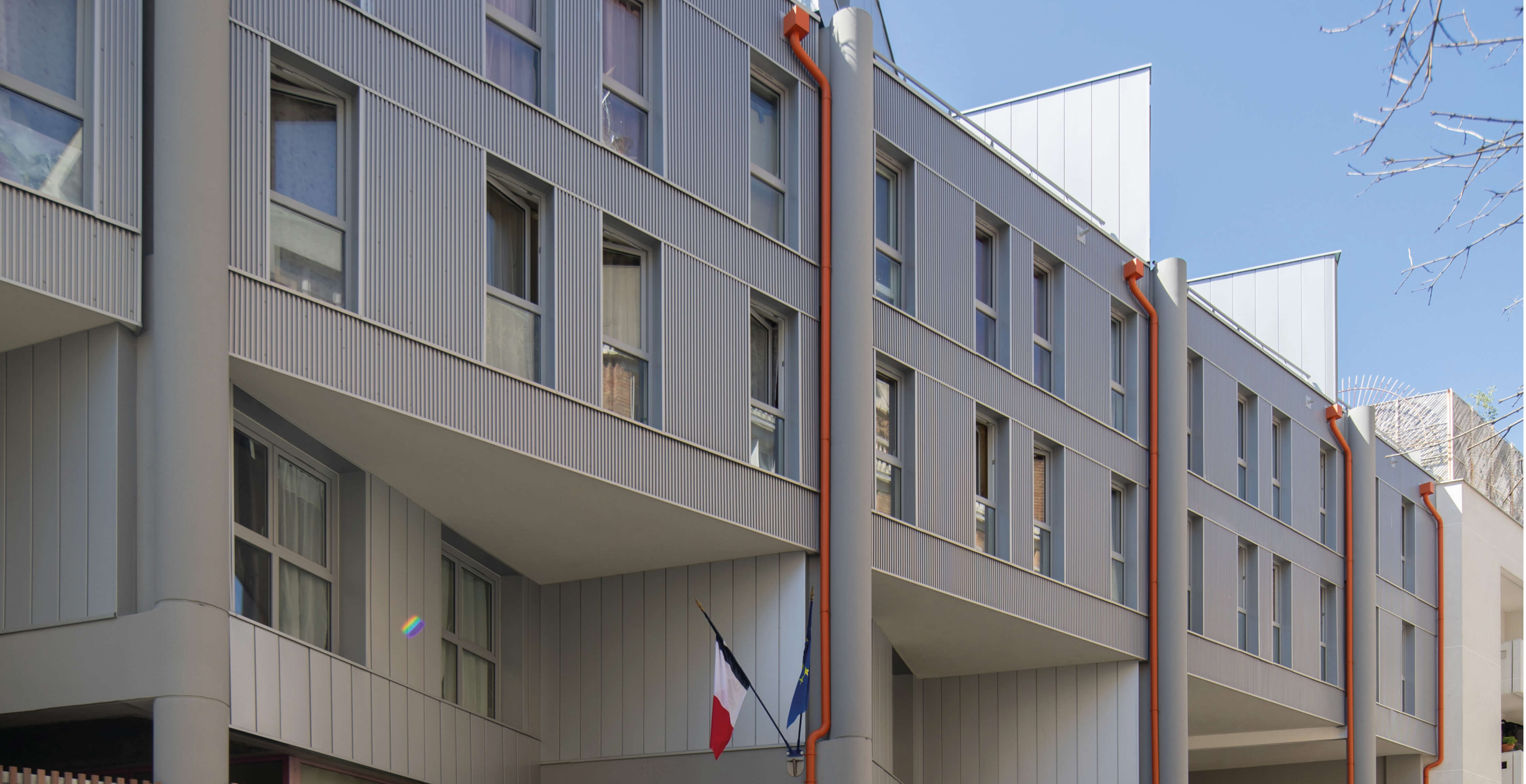 Photo avec ciel bleu en fond de la façade de la rue de Fontarabie avec drapeaux français et européen de l’école du rez-de-chaussée après la réhabilitation énergétique plan climat