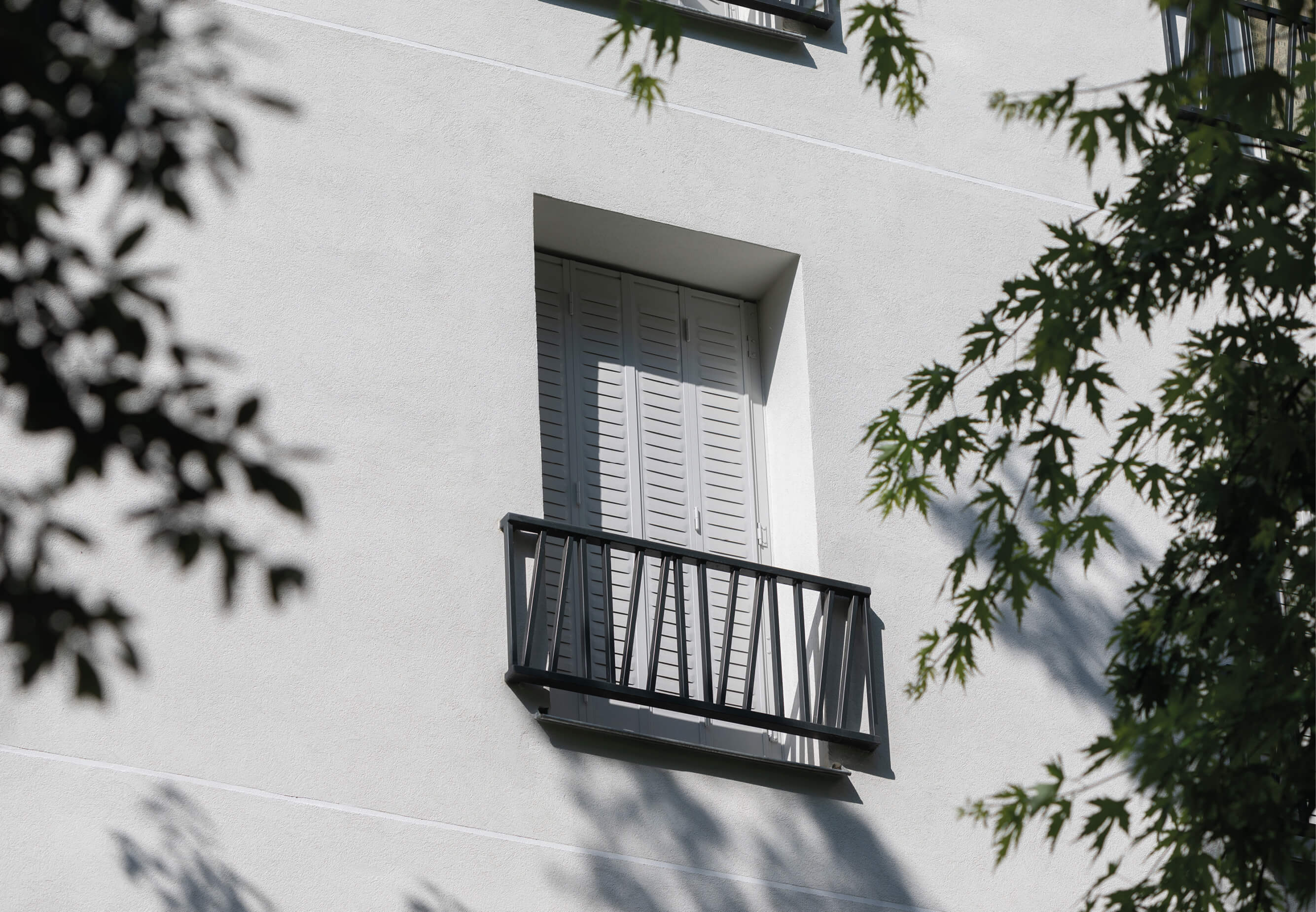 Photo d’une fenêtre volets fermés, baignée par le soleil, avec quelques branches d’arbre dans le cadre, sur la façade de l’immeuble du projet réhabilitation plan climat en milieu habité rue Jeanne d’Arc