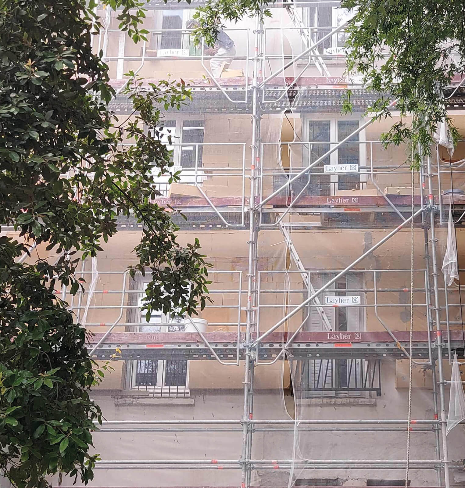 Photo d’une partie de l’échafaudage réhabilitation plan climat en milieu habité rue Jeanne d’Arc encadrée par les branches du jardin à l’arrière du bâtiment