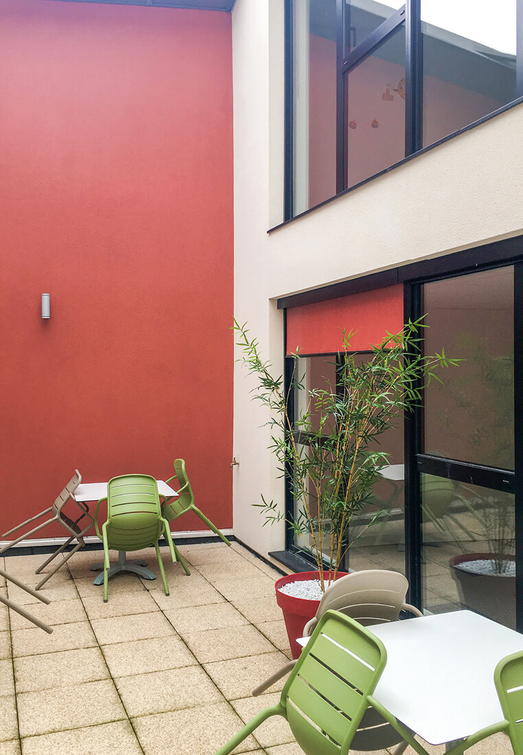 Photo d'un patio amenage avec des tables et chaises en metal vertes et beiges, une plante, avec un en fond mur rouge et sur le cote baie vitree