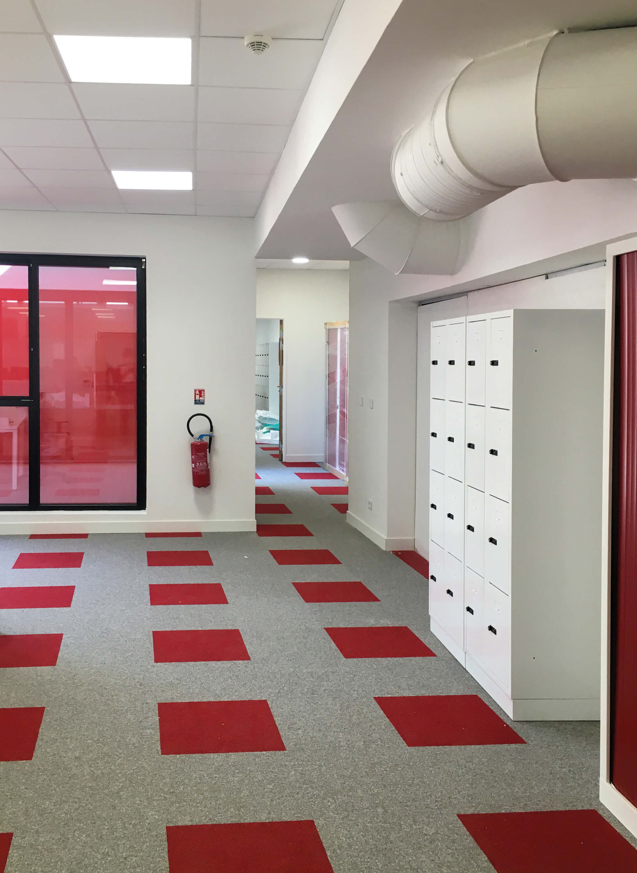 Photo d'un couloir avec moquette grise rehaussee de carres rouges et casiers de rangement.